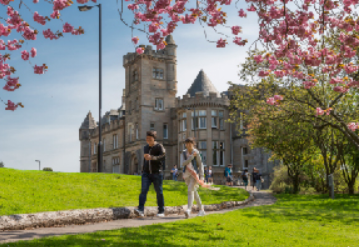 Image of Stirling campus - it is green, there are some trees with pink blossom, and a castle-like building in the background. Two students walk across the scene.